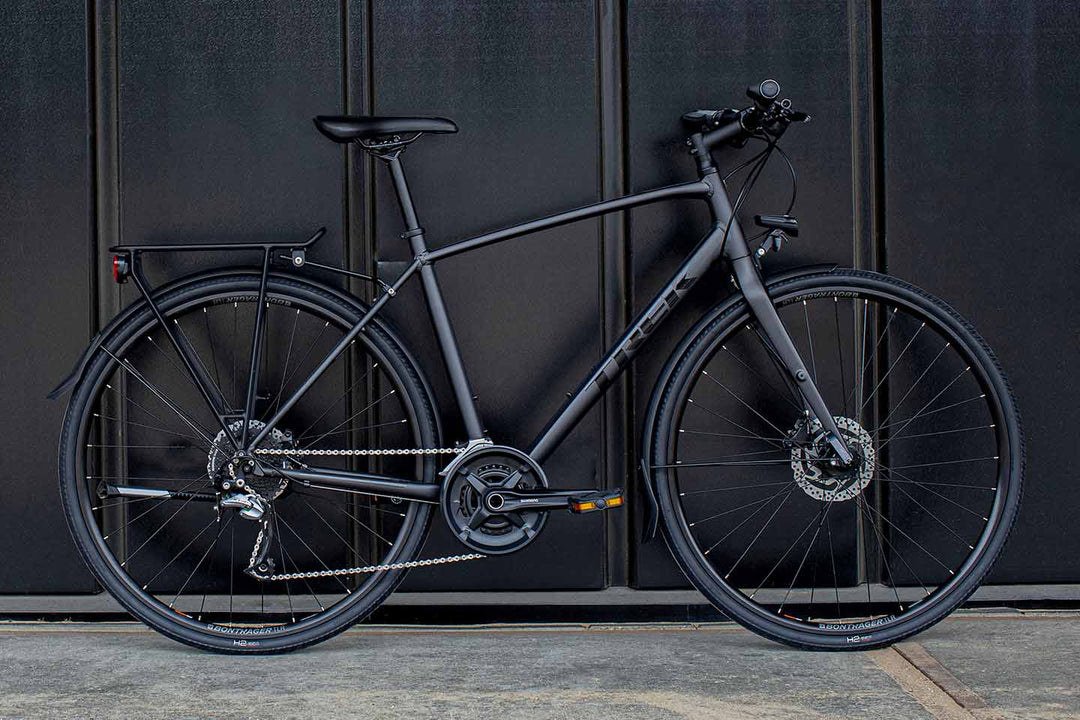 一面黒色の壁に立てかけたマットブラックのクロスバイクの写真。ライトやフェンダー、キャリアなどのアクセサリーも取り付けてある。