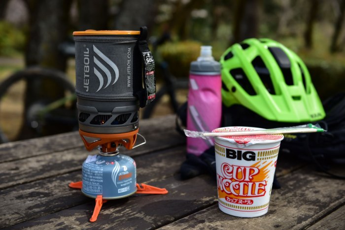 ジェットボイルとサイクルボトル、ヘルメット、そしてカップヌードルを屋外の木製テーブルの上に置いた写真